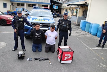 Dos policías con uniforme trasladan a dos hombres esposados frente a una patrulla, junto a armas, un casco y una mochila roja en el suelo