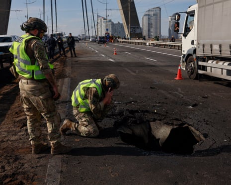 Soldiers in camouflage and hi-vis vests kneel beside a large hole on Kyiv's Northern Bridge after a drone strike
